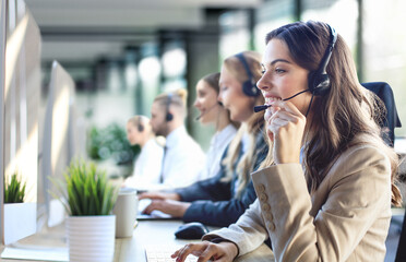 Female customer support operator with headset and smiling, with collegues at background. - 785936784
