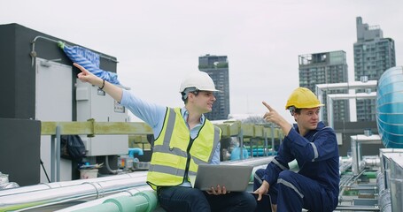 engineers manager and worker sitting on rooftop review plans on a laptop pointing to something at construction site with high-rise buildings