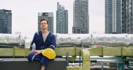 A engineer worker man in uniform sits on a bench and smile looking to camera at rooftop construction site. The scene takes place in a city with tall buildings in the background