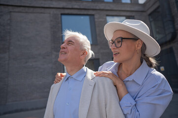 A woman in a hat and glasses hugs an elderly man in a white jacket from behind. Romantic relationships of mature people. 