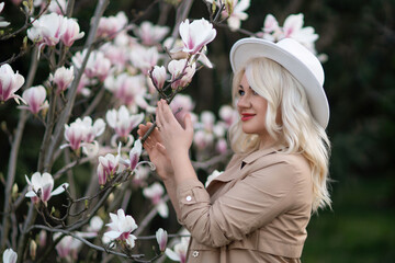 Naklejka premium Magnolia flowers woman. A blonde woman wearing a white hat stands in front of a tree with pink flowers. She has a smile on her face and she is enjoying the beautiful scenery.