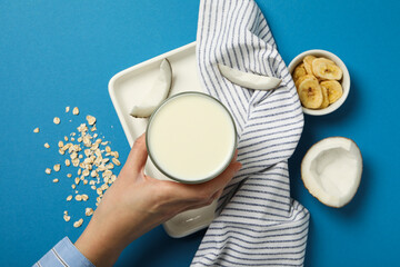 Glass of milk in hand, towel, coconut and sliced bananas on blue background, top view