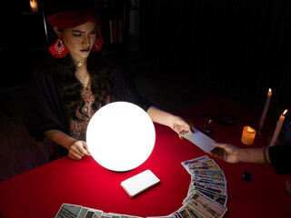 THAILAND - DEC 19, 2019: Asian tarot card reader in a headscarf focuses on a tarot deck with a crystal ball casting light over the table surrounded by candles in a mysterious black room
