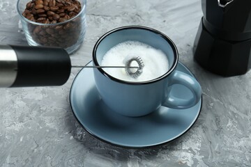 Whisking milk in cup with mini mixer (milk frother) on grey textured table, closeup