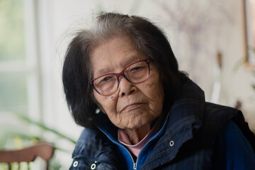 An elderly woman looks seriously at the camera. She is leaning to the side, with a warm vest on....