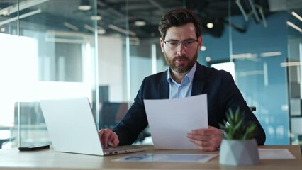Front view of cheerful businessman rewriting information from piece of important paper in wireless laptop in personal cabinet. Handsome bearded male engaging in paperwork with modern gadgets indoors.