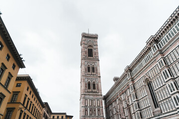 Fototapeta na wymiar Medieval buildings and Santa Maria del Fiore's Gothic-Renaissance intricate façade framing Giotto's Campanile from Piazza del Duomo, standing tall in the centre, against the cloudy sky.