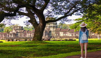 A beautiful woman in a white hat and mini jean shorts walks towards the temple - Acacia tree on the background of the temples - Green leaves pattern of acacia tree - Angkor temple complex, Siam Reap
