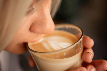 A blonde woman is drinking coffee from a glass. She is smiling and she is enjoying her drink.