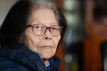 An elderly Asian woman turns to the camera and makes eye contact with a pensive expression. She is...