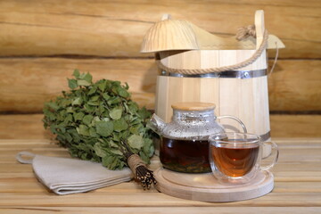 A glass cup of herbal tea stands on a wooden bench next to a dry birch broom and traditional sauna accessories in the interior of a log bathhouse. 