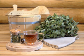 A glass cup of herbal tea stands on a wooden bench next to a dry birch broom and traditional sauna...