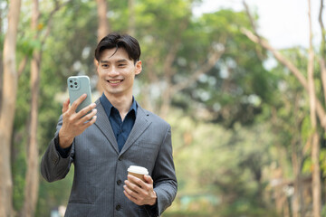 Confident asian businessman holding takeaway coffee cup using mobile phone in the park