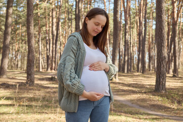 Adorable gentle brown haired Caucasian pregnant woman waking in spring forest wearing knitted jumper touching her belly breathing fresh air on last months of pregnancy