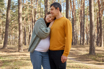 Fototapeta premium Beautiful young family walking in spring forest hugging and admiring beautiful forest pregnant woman leaning on husband shoulder enjoying happy moments