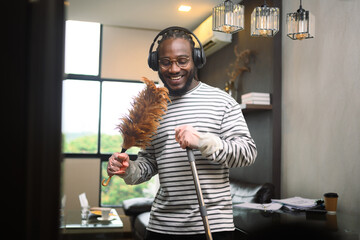Happy African American man having fun cleaning house and singing using duster as microphone.