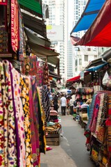 Naklejka premium A bustling textile market with bolts of colorful fabrics, intricate embroideries, and handmade garments on display, surrounded by towering skyscrapers and bustling streets, Generative AI