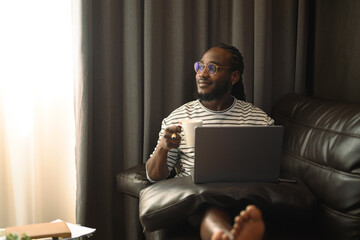 Positive African millennial man drinking coffee on couch and working with laptop