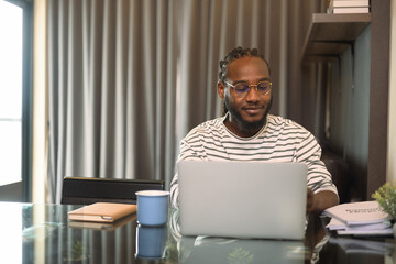 Smiling bearded African man working remotely from home using laptop on dinning table
