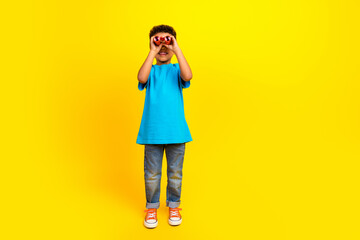 Full length photo of adorable small child with curly hair dressed blue t-shirt staring in binoculars isolated on yellow background