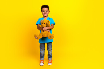 Full length photo of optimistic child with curly hair dressed blue t-shirt denim jeans cuddle teddy bear isolated on yellow background