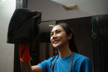 Beautiful female housekeeper in rubber gloves cleaning mirror with rag and spray in a modern bathroom