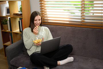 Relaxed young caucasian woman eating popcorn and watching movie on a laptop at home
