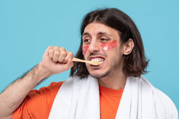 Studio shot of cheerful haired middle eastern man brushes teeth applies pink hydrogel patches under...