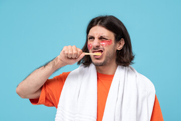 Studio shot of cheerful haired middle eastern man brushes teeth applies pink hydrogel patches under eyes with white towel on blue background. comic funny emotional