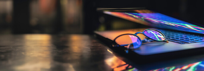 Glasses lie on the laptop, reflecting light from the screen in the dark.