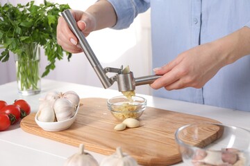 Woman squeezing garlic with press at white wooden table in kitchen, closeup