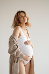 A beautiful pregnant girl in a white bodysuit and an oversized jacket, wavy hair. A pregnant beauty is expecting the birth of a child. Clothes for pregnant women, copy space. Motherhood and Childhood