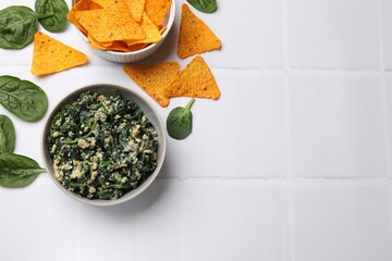 Tasty spinach dip with egg in bowl and nachos chips on white tiled table, flat lay. Space for text