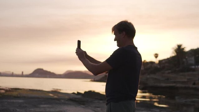 Handsome man taking panoramic photo on phone of rocks and sea in setting sun