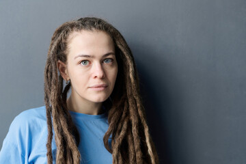 Minimal portrait of young Caucasian woman with long dreadlocks looking at camera standing on grey...