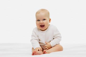 Cheerful baby with sparkling blue eyes and joyful smile sits on white bed, dressed in snug, white long-sleeved top, radiating happiness and innocence. Concept of childhood, motherhood, life, birth. Ad