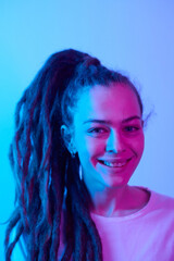 Vertical portrait of smiling Caucasian woman with long dreadlocks hairstyle in ponytail looking at camera in blue neon light