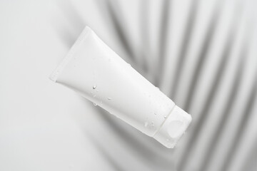 White body or face cream tube mockup with water drops on gray background with palm branch shadow. Skin moisturizing beauty cosmetics concept.