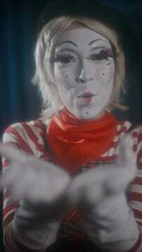 Vertical shot of romantic mime artist showing heart with hands, blowing kiss and smiling on camera during funny performance on stage