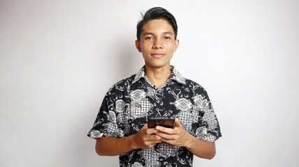 Happy young handsome Asian man wearing batik shirt posing holding smartphone