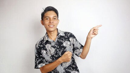 Happy young handsome Asian man wearing batik shirt posing pointing to the side towards open copy...