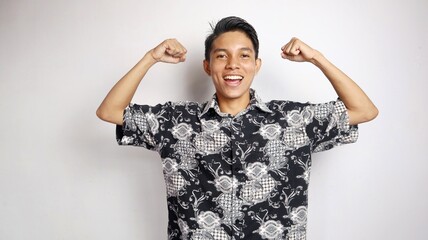 happy young handsome asian man wearing batik shirt posing raising hands excitedly,...