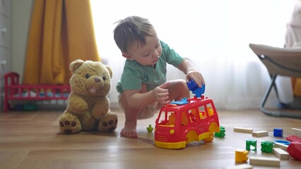 baby boy plays with toy bear a toy bus and puts wooden sticks in the hatch. development of fine motor skills concept. baby learns to put sticks toys into a bus car indoors in dream kindergarten