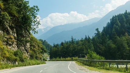 Scenic shot of a road surrounded by mountains, green grass, and trees on a sunny day