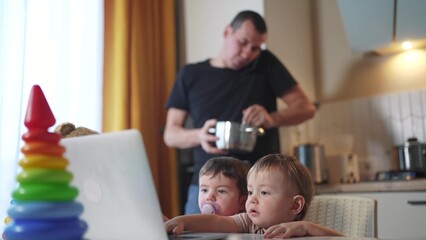 father cooking in the kitchen remote work. baby twins playing with laptop in the kitchen. father...