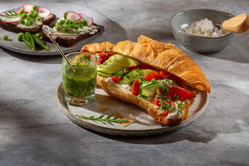 Croissant sandwich with cream cheese, smoked salmon, cucumber and arugula served with pesto on gray background.