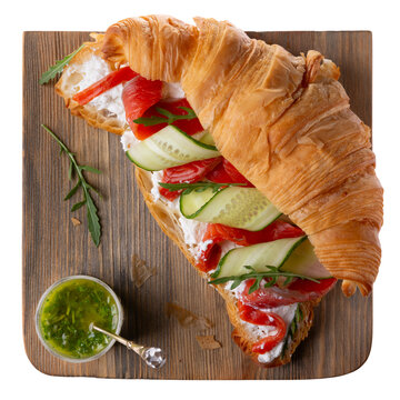Croissant sandwich with cream cheese, smoked salmon, cucumber and arugula served with pesto on wooden board isolated on white. Top view.