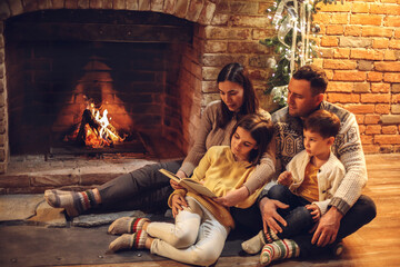 Happy european family with children sitting on floor by fireplace reading and frying marshmallow...