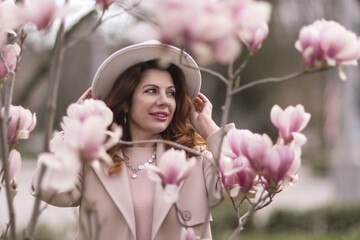 Fototapeta premium Woman magnolia flowers, surrounded by blossoming trees., hair down, white hat, wearing a light coat. Captured during spring, showcasing natural beauty and seasonal change.