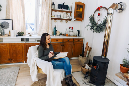 Back view photo of woman sitting near potbelly stove at home, female warming hands after winter walk in frosty weather, New Years home decor around. People and winter holidays concept